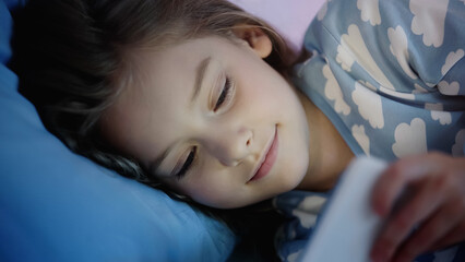 Smiling preteen child looking at blurred smartphone on bed.