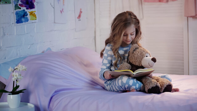 Smiling kid in pajama reading fairytale near soft toy on bed in evening.