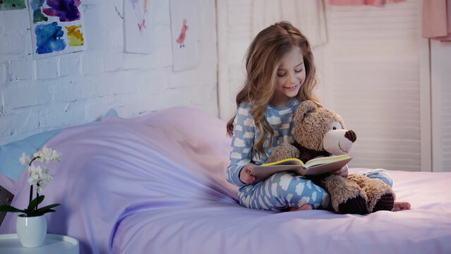 Smiling preteen child in pajama reading book near soft toy on bed in evening.