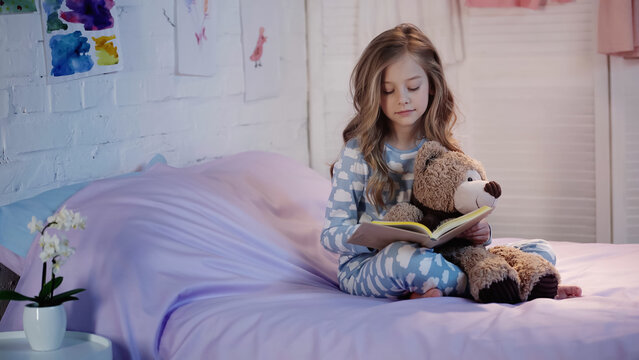 Preteen kid holding soft toy and reading book on bed.