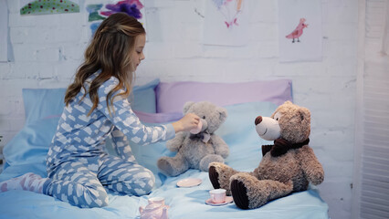 Side view of preteen kid in pajama holding cup near soft toy on bed.