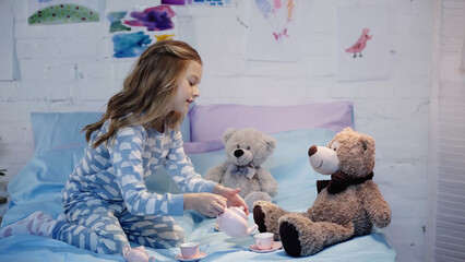 Side view of smiling preteen kid pouring tea near soft toys on bed.