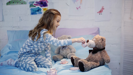 Side view of girl in pajama holding cup near teddy bear on bed.