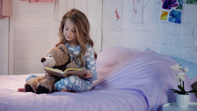 Preteen child in pajama reading book near teddy bear in bedroom.