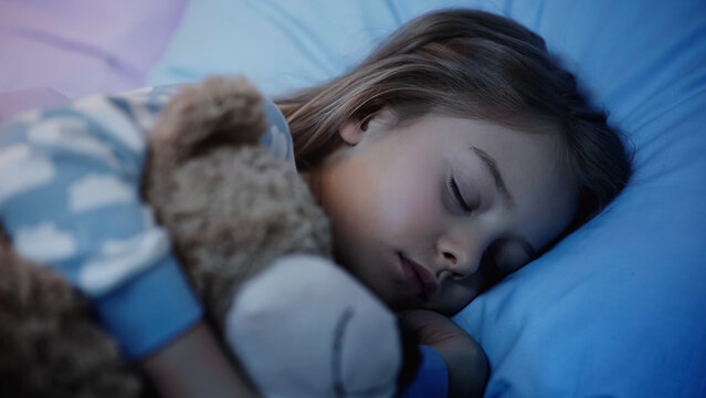 Preteen girl sleeping near blurred soft toy on bed at night.
