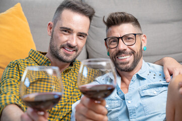 Gay couple in love sitting at home on the floor and toasting with red wine. High quality photography.