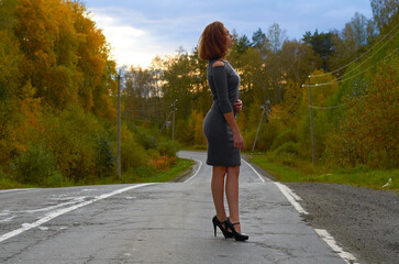 Fototapeta premium a young girl in a gray dress and black shoes with a backpack on her back, walking along an old highway against the background of a colorful forest and sunset