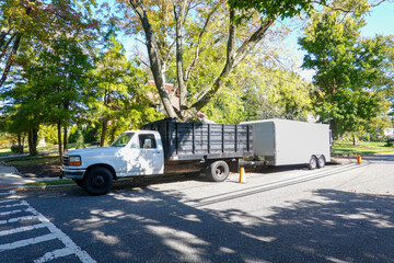A white landscaping truck with a long white enclosed trailer trailer seen on a shady residential asphalt street