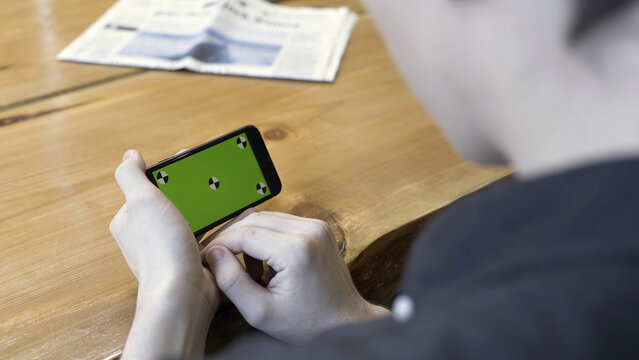 Smartphone Horizontal Orientation With A Green Screen: Shot Over The Shoulder Of A Young Man. Stock Footage. Man Sitting At The Table, Tapping And Scrolling On The Chromakey Of The Phone.