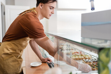 Side view of young seller in glove taking dessert from showcase in sweet shop.