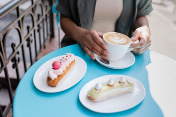 Cropped view of african american woman holding cup of cappuccino near eclairs on terrace of sweet shop.