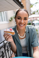 Young african american woman holding dessert and looking at camera on terrace of sweet shop.