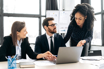 Multiracial colleagues are brainstorming on strategy together, sitting in modern creative office, studying documents, using laptop, discussing. Team collaboration of business people