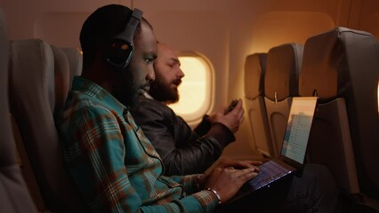 African american freelancer using laptop on flight, travelling on work trip during sunset in economy class. Working on computer and flying on airplane, aviation journey. Handheld shot.
