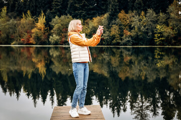 Fototapeta na wymiar Happy young pretty woman walking outdoors in autumn park using mobile phone chatting, scenic view of the river
