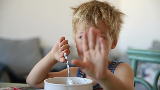 Portrait of cute adorable little blond caucasian sleepy kid boy eating oatmeal porridge early morning. Grumpy child showing stop enough gesture raising hand. Children healthy food nutrition