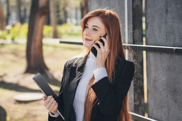 Ginger businesswoman with laptop making call by phone outdoors while walking through the city park. Business communication. Business portrait. Online education.
