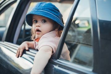 Baby girl sticking her head out of a car window, looking back as he and his family go on a journey. Summer road trip. Traveling with children.