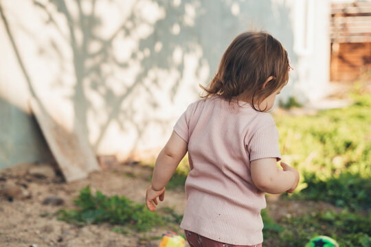 Rear View Of A Baby Girl Running Through The Yard Of The House And Playing. Baby Development. Back View.