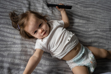 One small caucasian girl toddler lying in diapers on the bed at home top view hold remote...