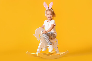 Cute little girl in bunny ears with rocking horse on yellow background
