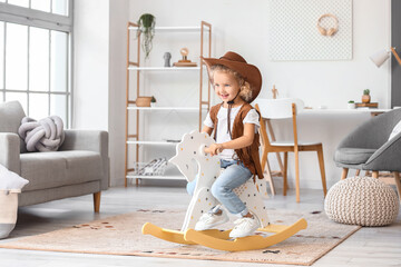 Cute little girl in hat and vest playing with rocking horse at home