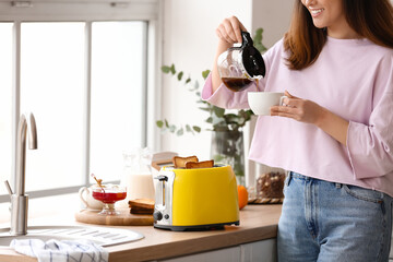 Beautiful young Asian woman drinking coffee and making tasty toasts in kitchen