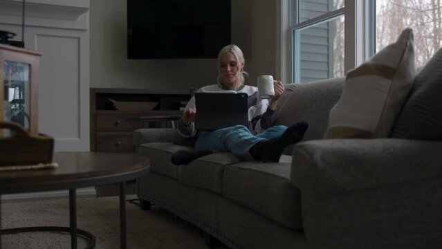 Woman sitting on the couch in front of a bay window, drinking coffee while scrolling on a tablet from far away and the side.
