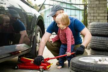 Cute toddler boy helping his father to change car wheels at their backyard. Father teaching his little son to use tools.