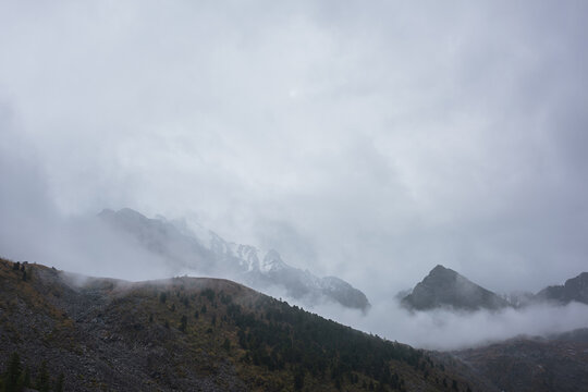 Dark atmospheric landscape with high mountain silhouettes in dense fog in rainy weather. Snowy rocky mountain top above hills in thick fog in dramatic overcast. Black rocks in low clouds during rain.