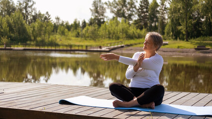 An elderly Caucasian, gray-haired woman does stretching exercises in the park on a yoga mat....