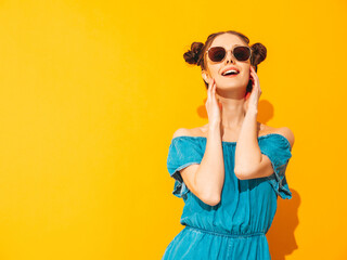 Young beautiful smiling female in trendy summer jeans overalls. Sexy carefree woman with two horns hairstyle posing near yellow wall in studio. Positive model having fun. In sunglasses. Isolated