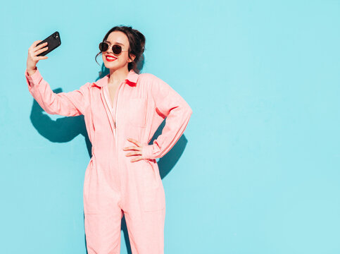 Young beautiful smiling female in trendy summer pink overalls. Sexy carefree woman posing near blue wall in studio with two horns. Positive model having fun and going crazy. Taking selfie photos