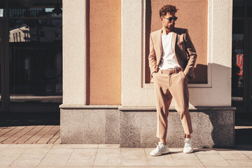 Handsome confident stylish hipster lambersexual model.Sexy modern man dressed in elegant beige suit. Fashion male posing in the street background in Europe city at sunset. In sunglasses. Full length - obrazy, fototapety, plakaty