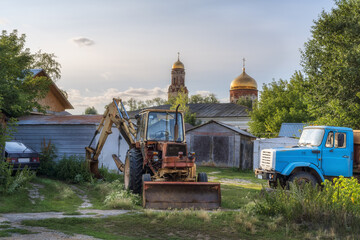 Rural courtyard with a tractor-loader, an old truck and a car and the golden domes of the church. Summer. Ural, Russia.