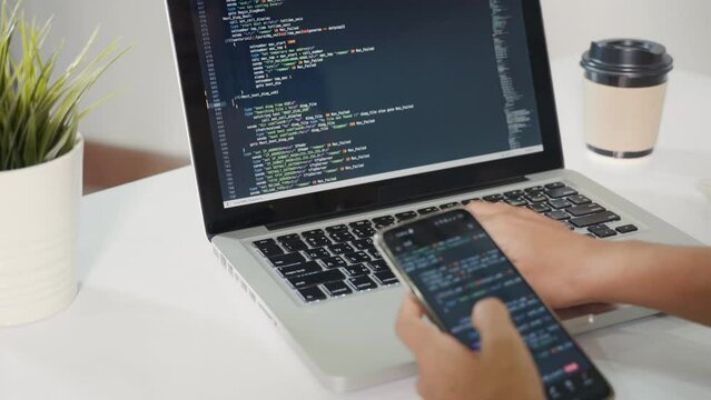 Programmer woman looking mobile phone program code at workplace, software developer hands typing data coding programming javascript with laptop computer, IT startup concept, Web application design