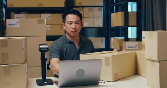 Asian businessman use barcode machine scan customer data in paper box and type register online information order detail in laptop for send express delivery at warehouse. Startup business concept.