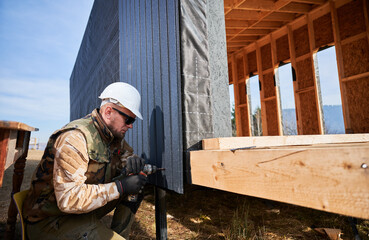 Male builder installing black corrugated iron sheet used as facade of future cottage. Close up of man worker building wooden frame house. Carpentry and construction concept.