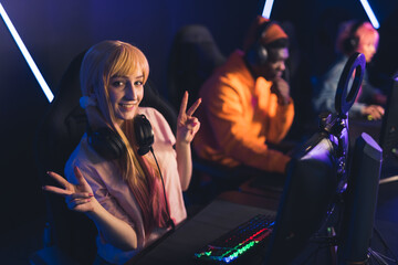 Teenage gamer girl wearing cute ponytails and headset sitting in gaming chair by professional computer smiling looking into camera showing peace signs. High quality photo