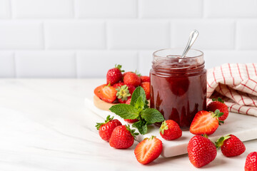 Strawberry jam in glass jar at white table.