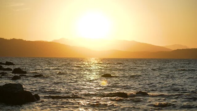 4k stock video footage of beautiful sunset on tropical beach. Natural sunny sunset background. Sea waves splashing, black silhouettes of mountains isolated on bright sunny orange sunset sky background