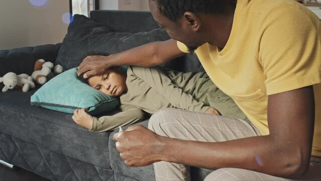 Medium long side view of young African American man sitting on couch, measuring body temperature of sick son
