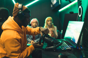 Black teenage guy wearing headset playing racing game with steering wheel watched by two teenage...