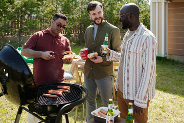 Three happy young intercultural men having beer and chat during outdoor party on weekend while one of them making barbecue