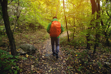 A tourist in a green jacket and a red backpack walks along a mountain path in spring or autumn. Traveler, tourist in autumn landscape. Mountains and trekking, rear view