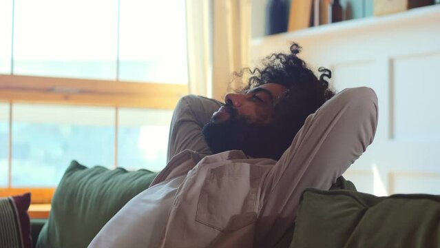 Young tired self satisfied Arabian man falls on sofa leaning back puts hands under head rejoicing at end difficult day and successful completion many different things sits in living room of house