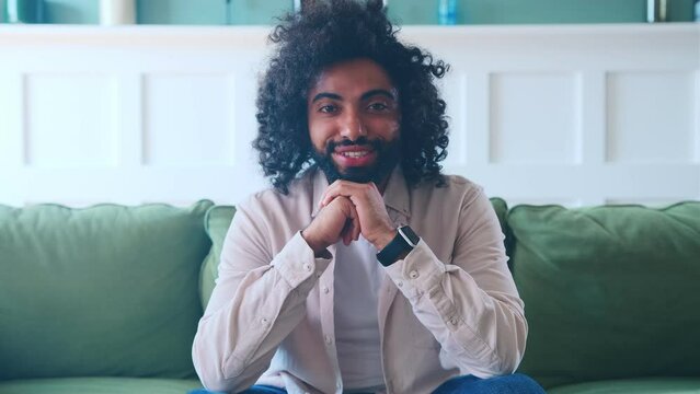 Young happy brunette middle eastern man student age begins to smile modestly looking straight at you and propping head with hands dressed in casual clothes sits on sofa in home living room.