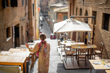 Sylish woman walks on narrow and cozy street in old town of Siena city. Concept of traveling in Tuscany region and italian lifestyle - obrazy, fototapety, plakaty