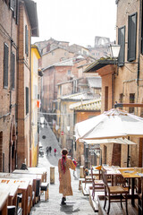 Fototapeta na wymiar Sylish woman walks on narrow and cozy street in old town of Siena city. Concept of traveling in Tuscany region and italian lifestyle