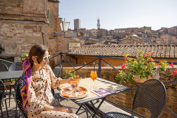 Fototapeta premium Young woman having lunch with pizza and wine at outdoor restaurant with beautiful view on the old town of Siena. Concept of italian cuisine and traveling Tuscany region of Italy